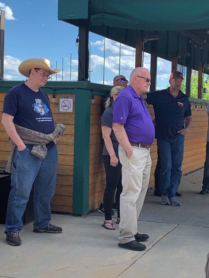 Group of people standing outside a wooden structure; man in cowboy hat and man in purple shirt are prominent. Cloudy sky.