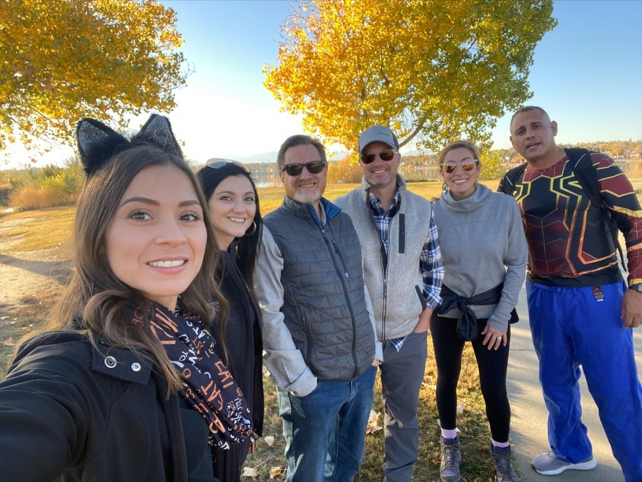 Group of people smiling, posing for selfie outdoors on a sunny day with fall foliage.