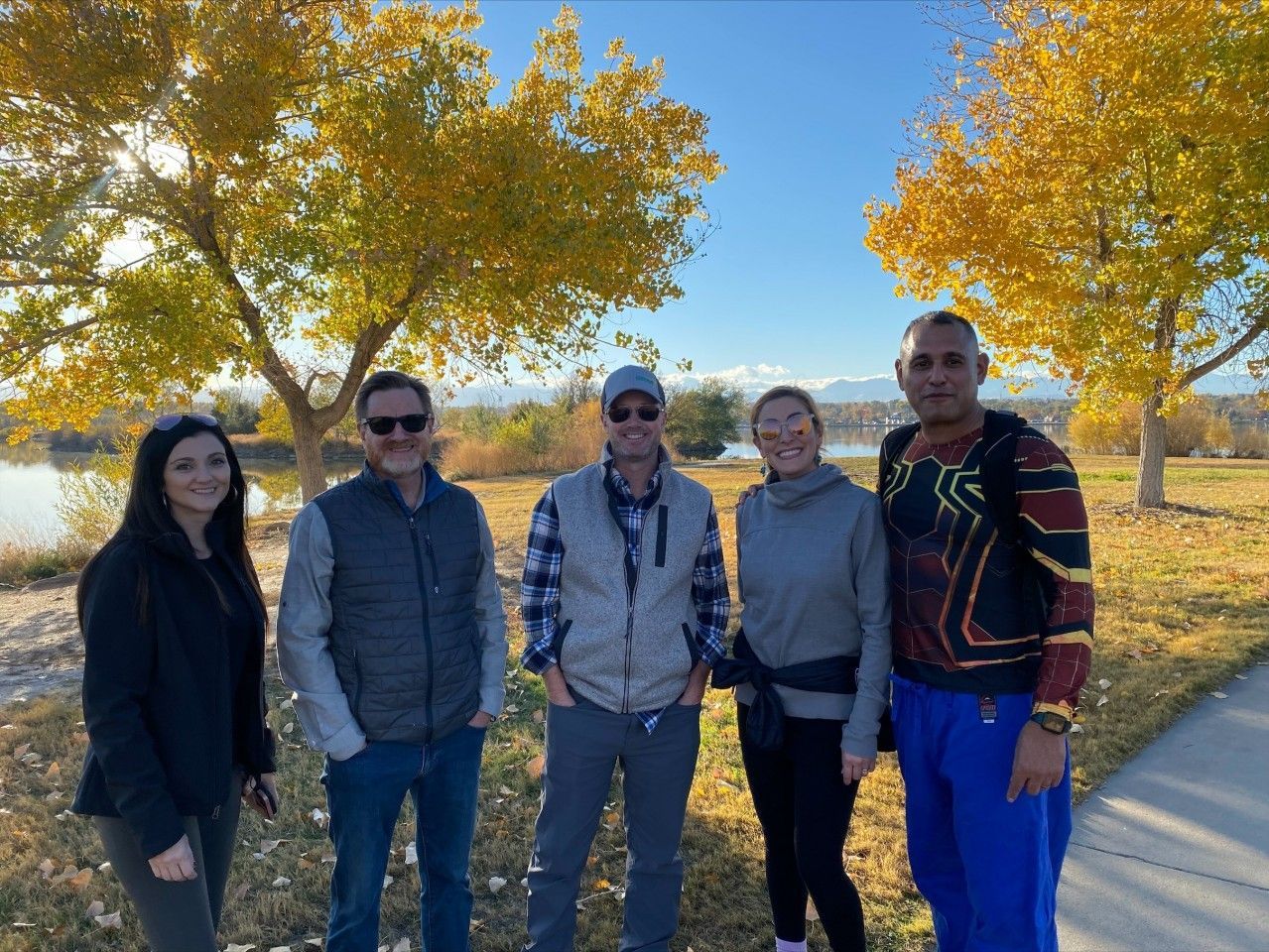Five people standing near a path, golden trees, water, and mountains in the background under a blue sky.