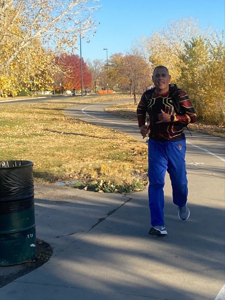 Man running on paved path, fall foliage in background. He's wearing blue pants and a patterned shirt.