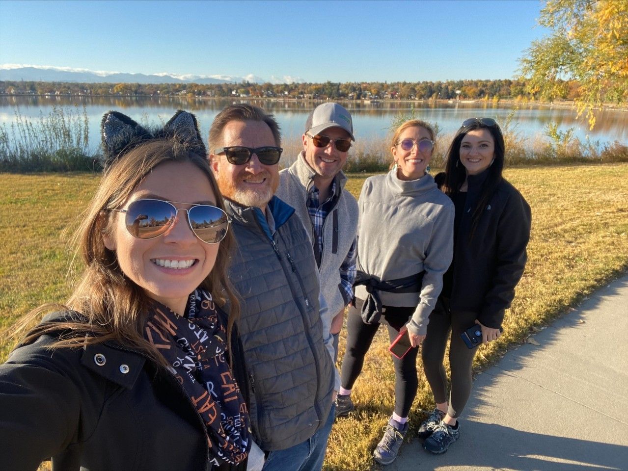 Five people smile for a selfie near a lake with fall foliage.