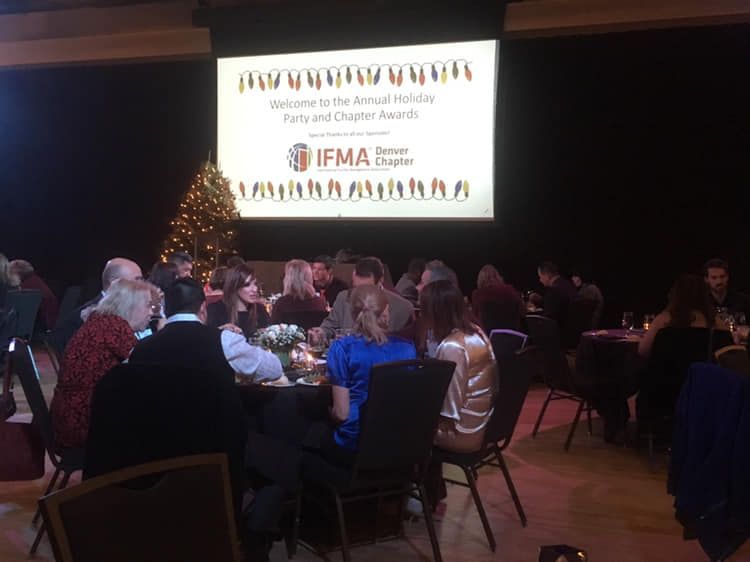People seated at tables during an awards event. A screen displays the event's title and logo.