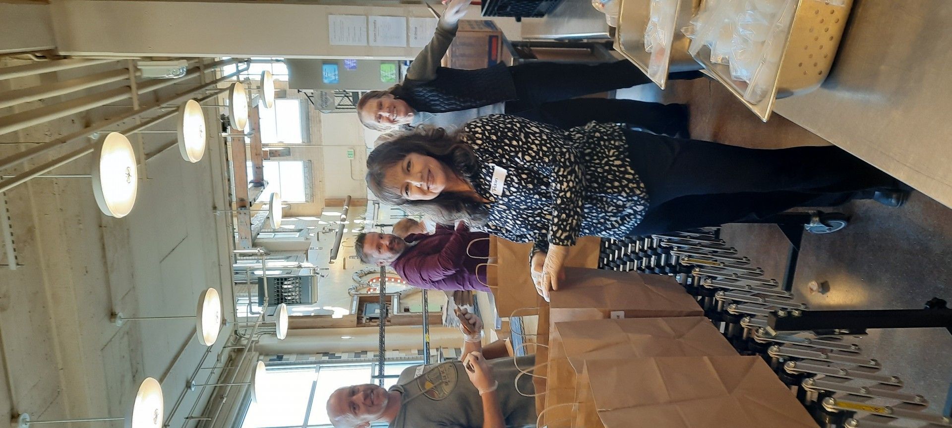 People packing boxes in a brightly lit room. A woman in a black and white patterned shirt smiles at the camera.