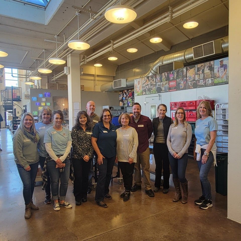 Group of people standing indoors, likely in a business. They smile, wear casual clothing, and stand under lights.
