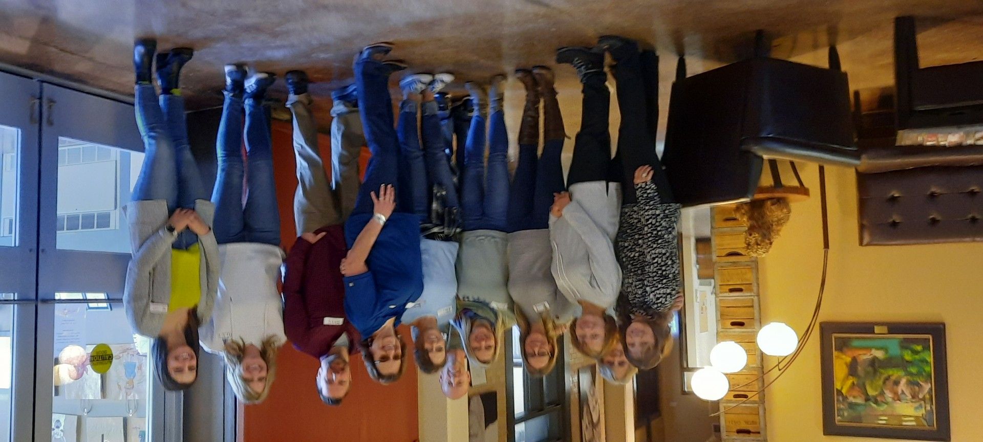 A group of people standing, smiling near a glass door in a building. They are wearing casual clothes.