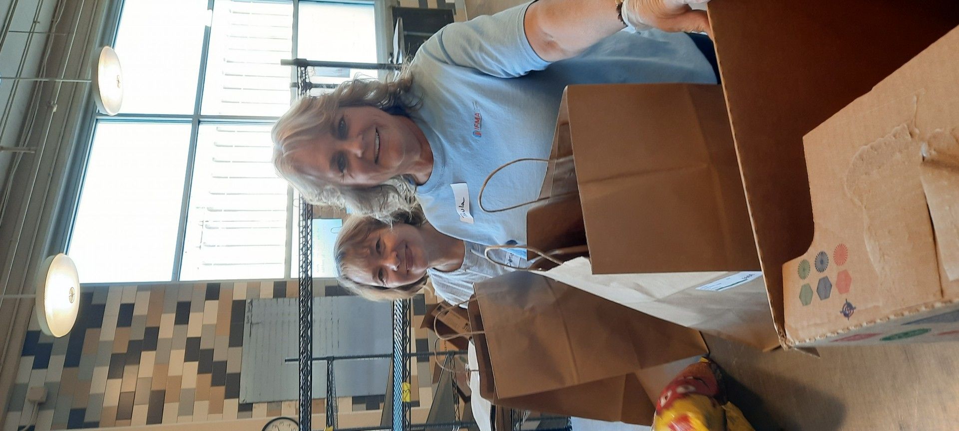 Two women packing items into brown paper bags and cardboard boxes in a well-lit room.