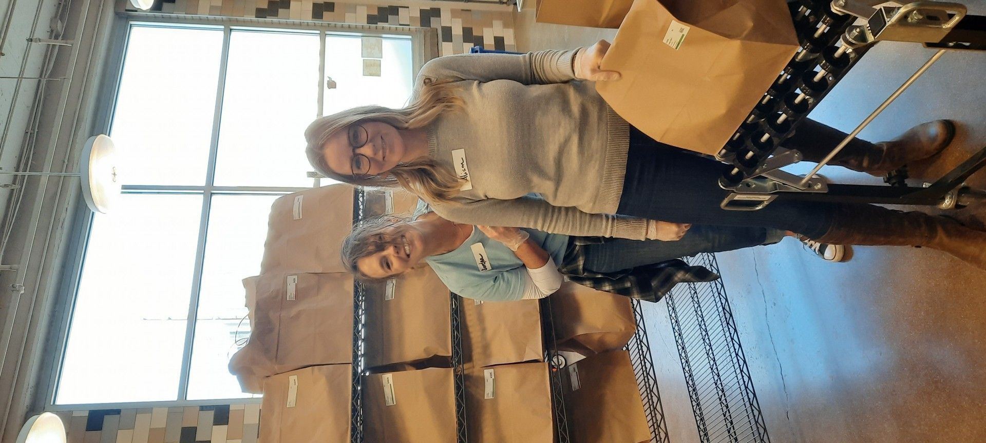 Two people stand near a shelf of boxes and hold bags. They appear to be in a warehouse setting.