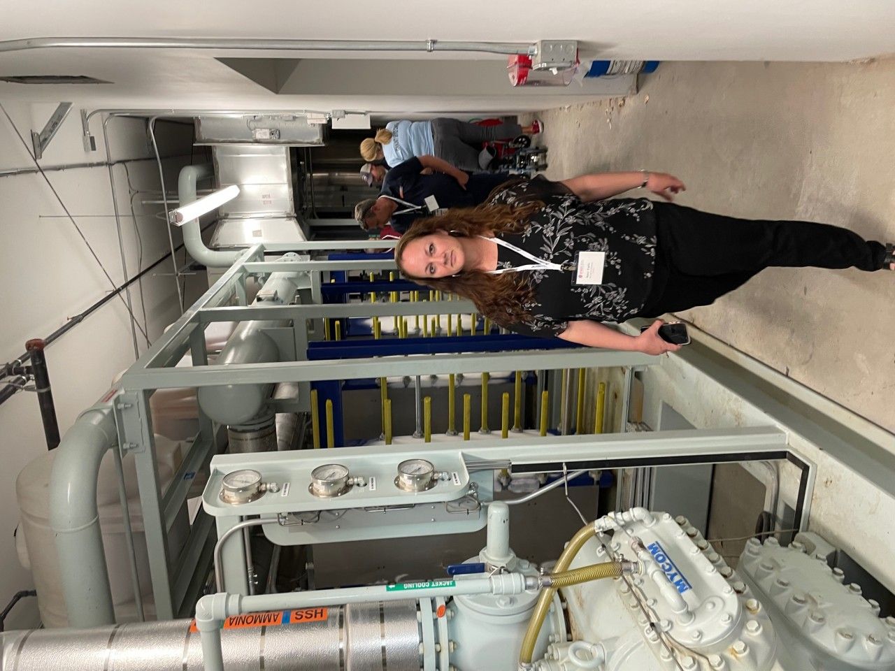 Woman stands in an industrial setting, near machinery and pipes, with others in the background.