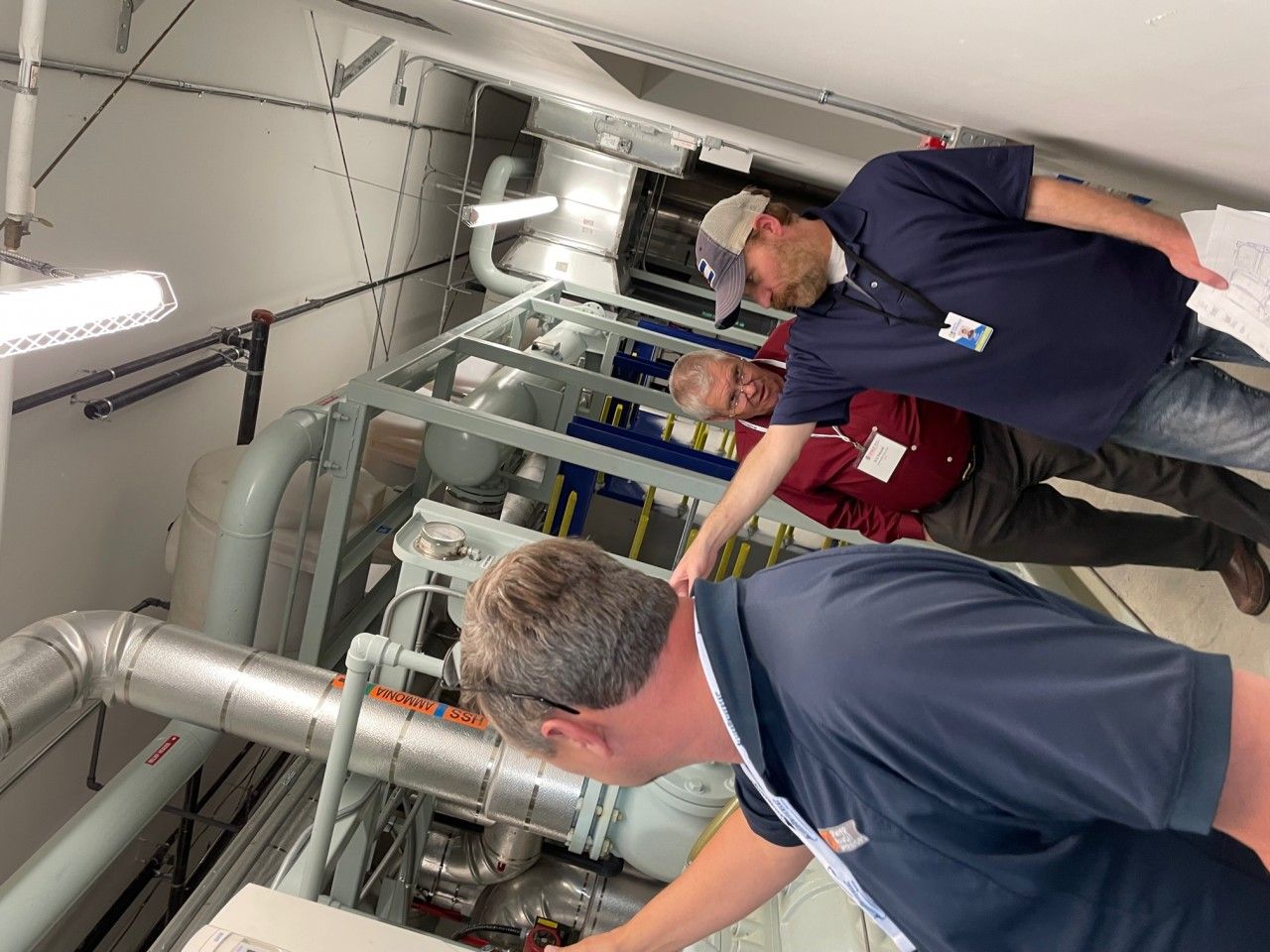 Three men inspecting equipment in a mechanical room, pointing at parts. Gray pipes, blue and red accents.