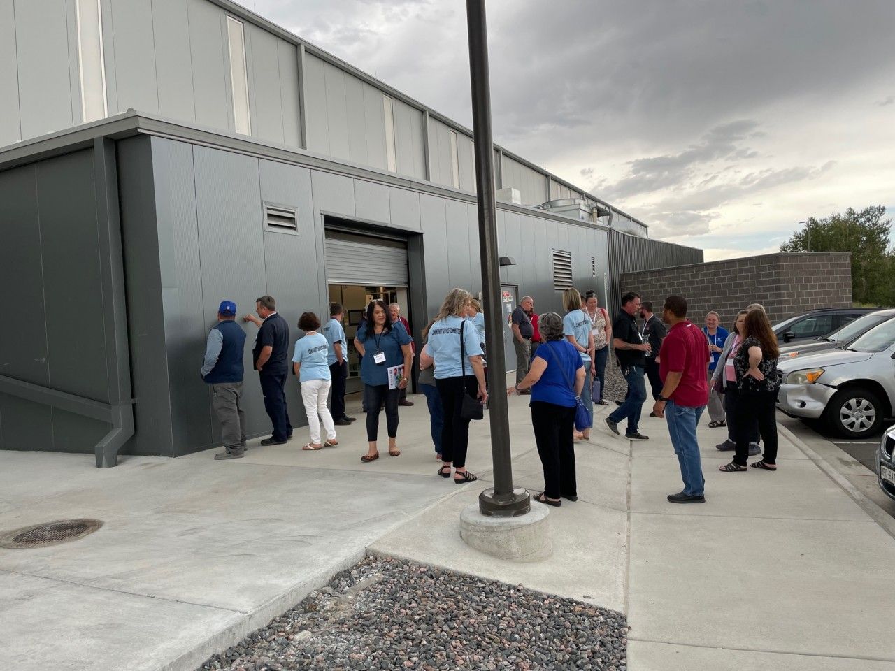 People gather outside a gray building with an open garage door, cloudy sky.