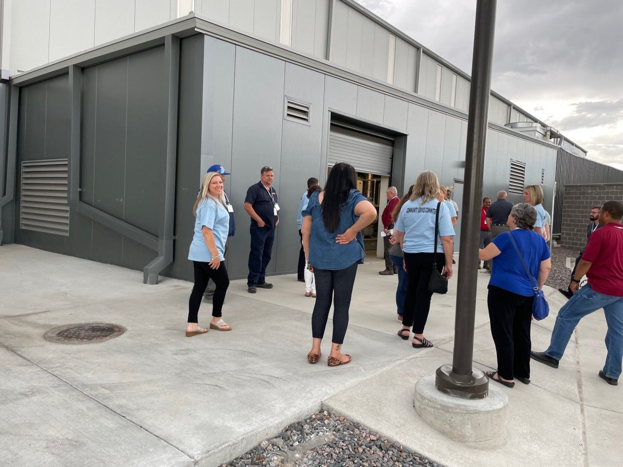 Group of people outside a gray building with an open door. Some wear blue shirts, standing on concrete.
