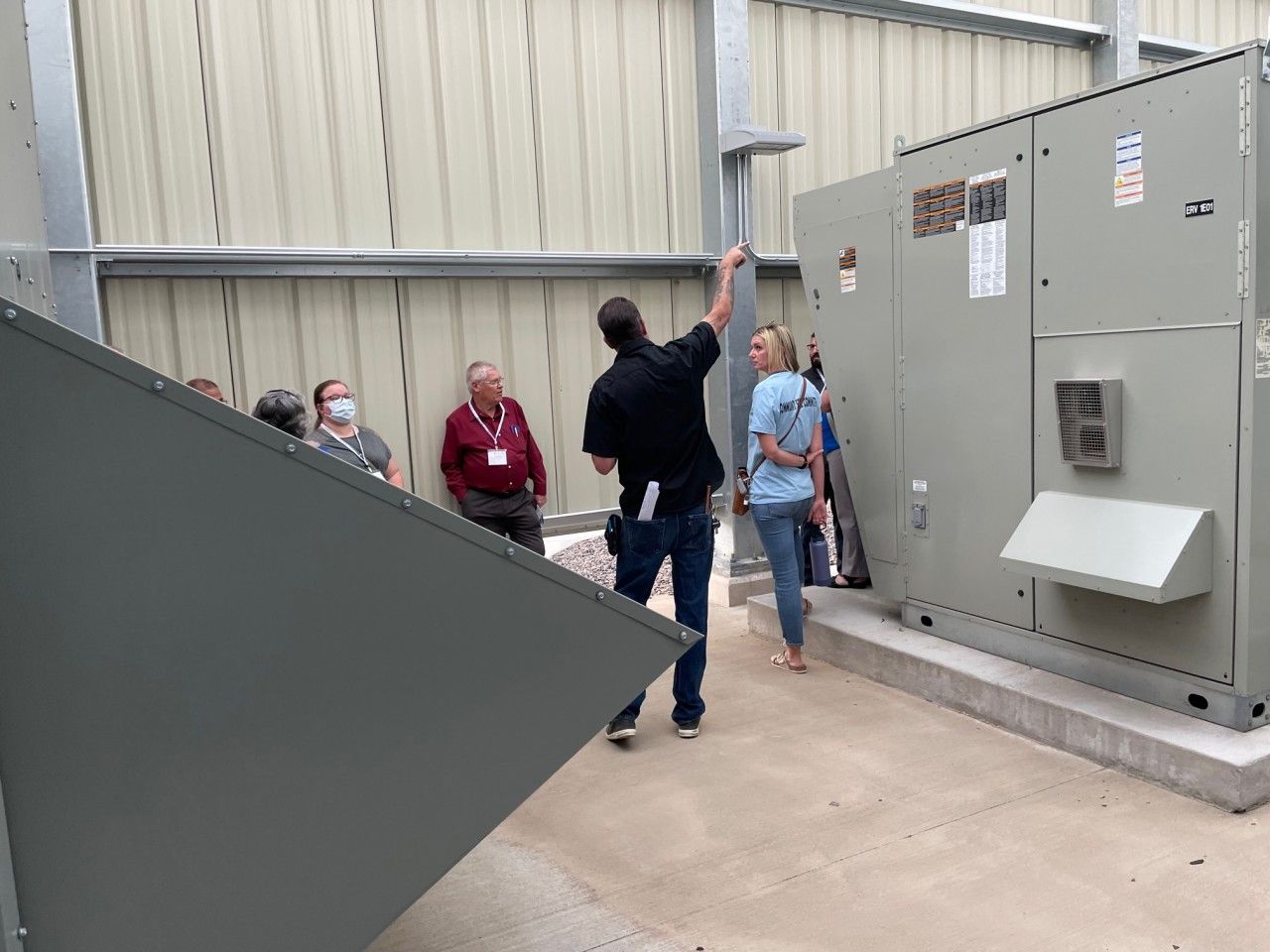 Group observes HVAC unit; man points, others watch. Building interior with gray equipment and walls.