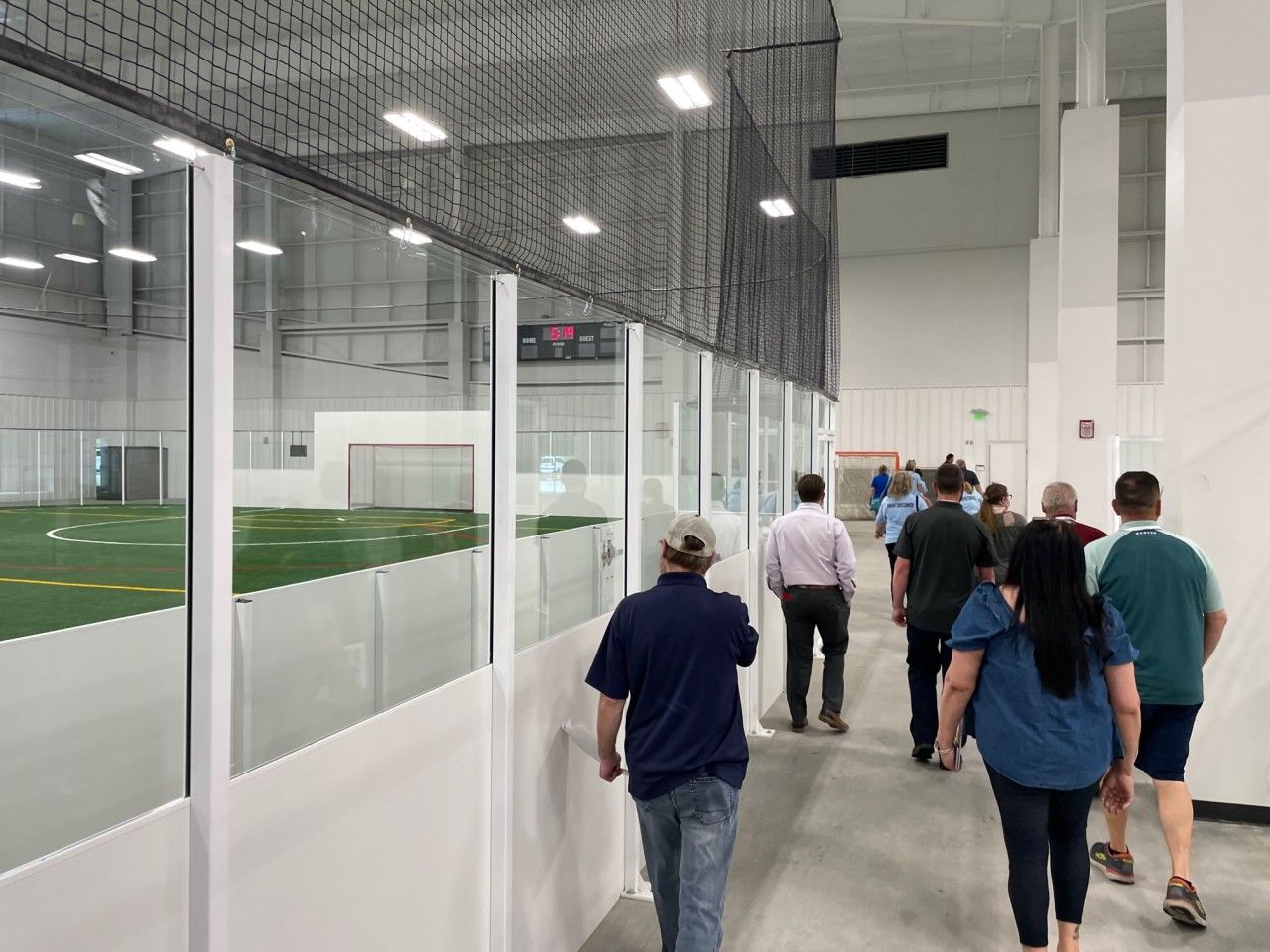 People walking down a hallway, looking into a sports field through glass windows.