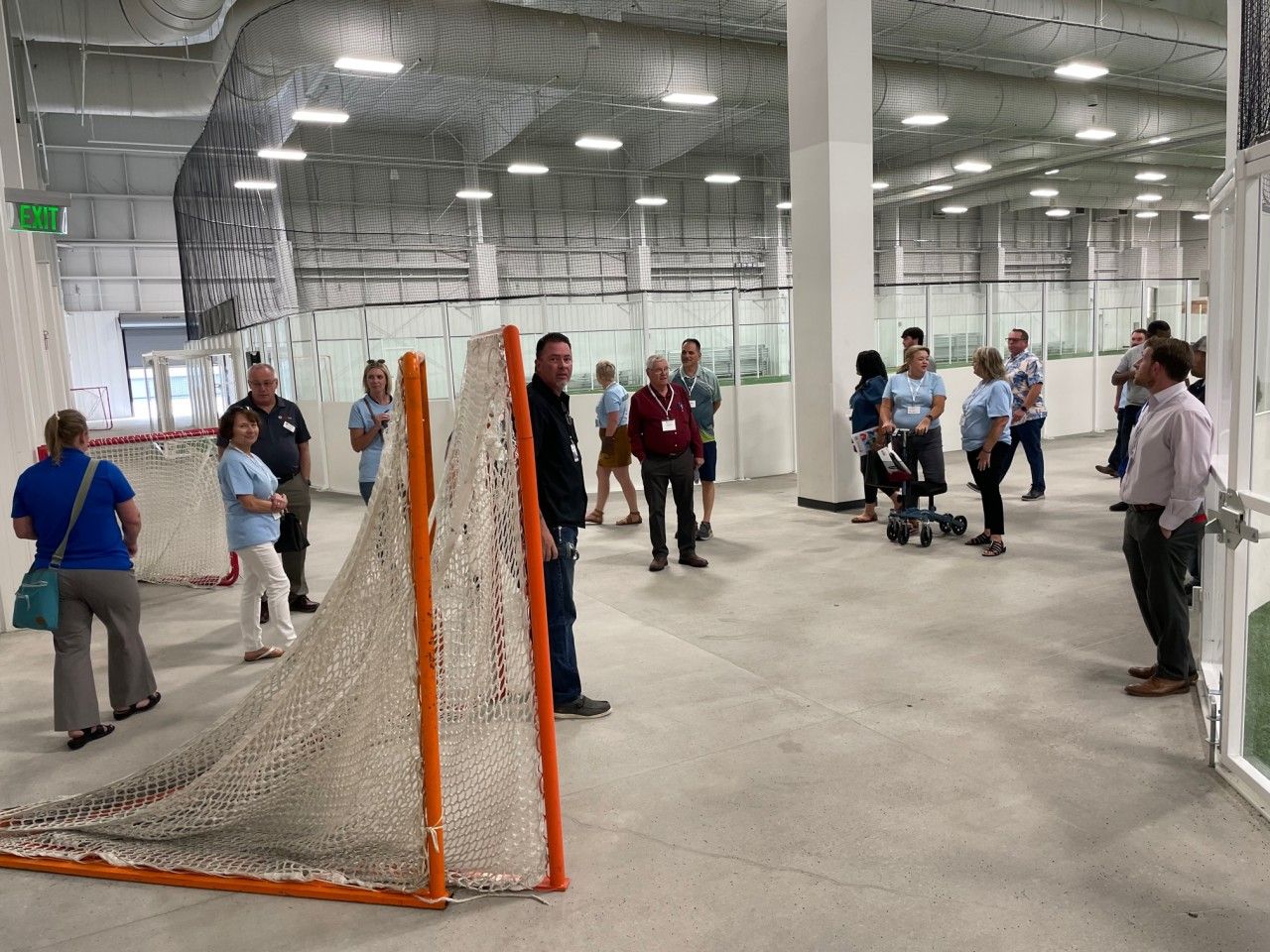 People in a sports arena, observing a lacrosse net. White, gray, and orange dominate the space.