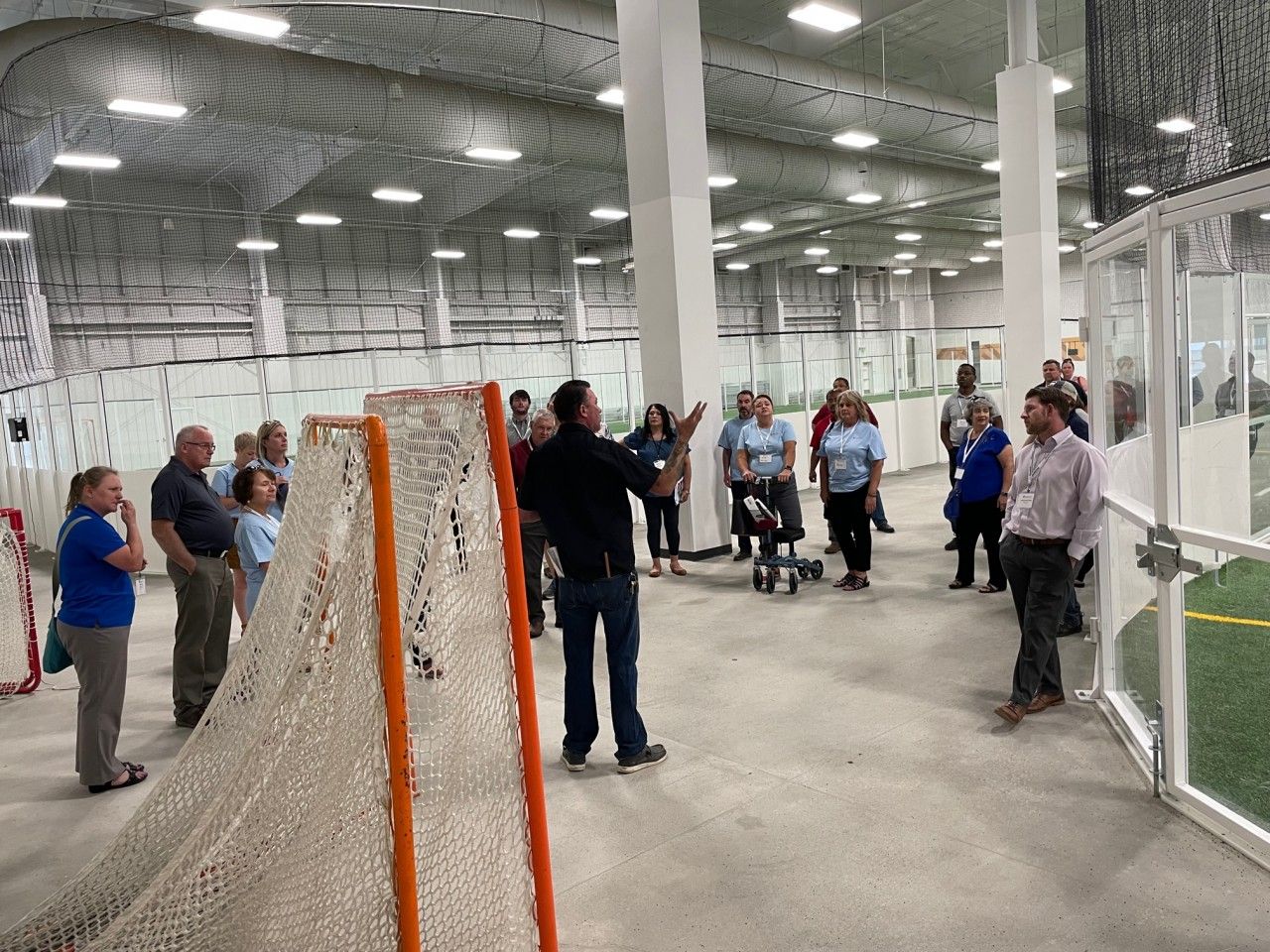 Indoor sports facility. Man gesturing to a group of people near a goal. Bright lighting, large open space, turf field.