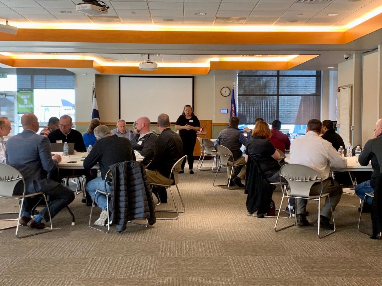 Group of people in a meeting room, a presenter standing near a screen.