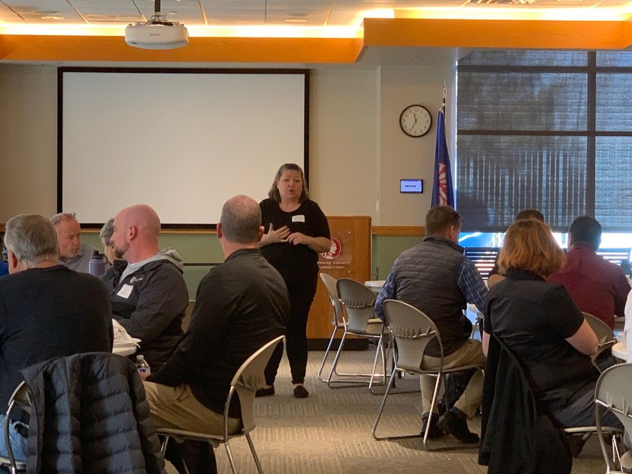 A woman presents to a seated audience in a conference room with a screen and flag.