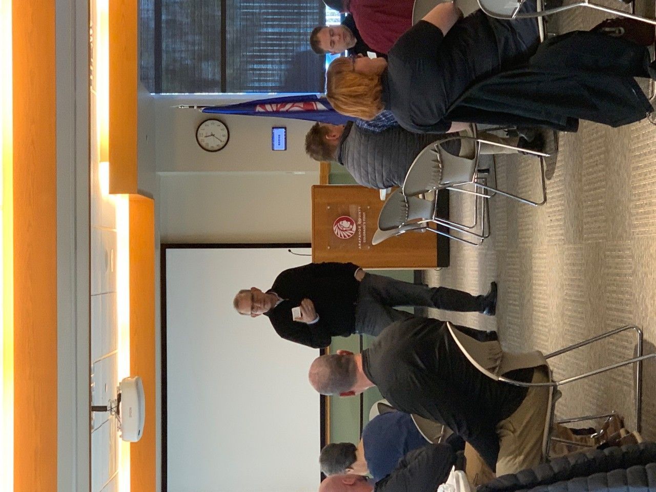 Man speaking at a conference. Others seated, watching. Projection screen, flag, clock. Orange wall.
