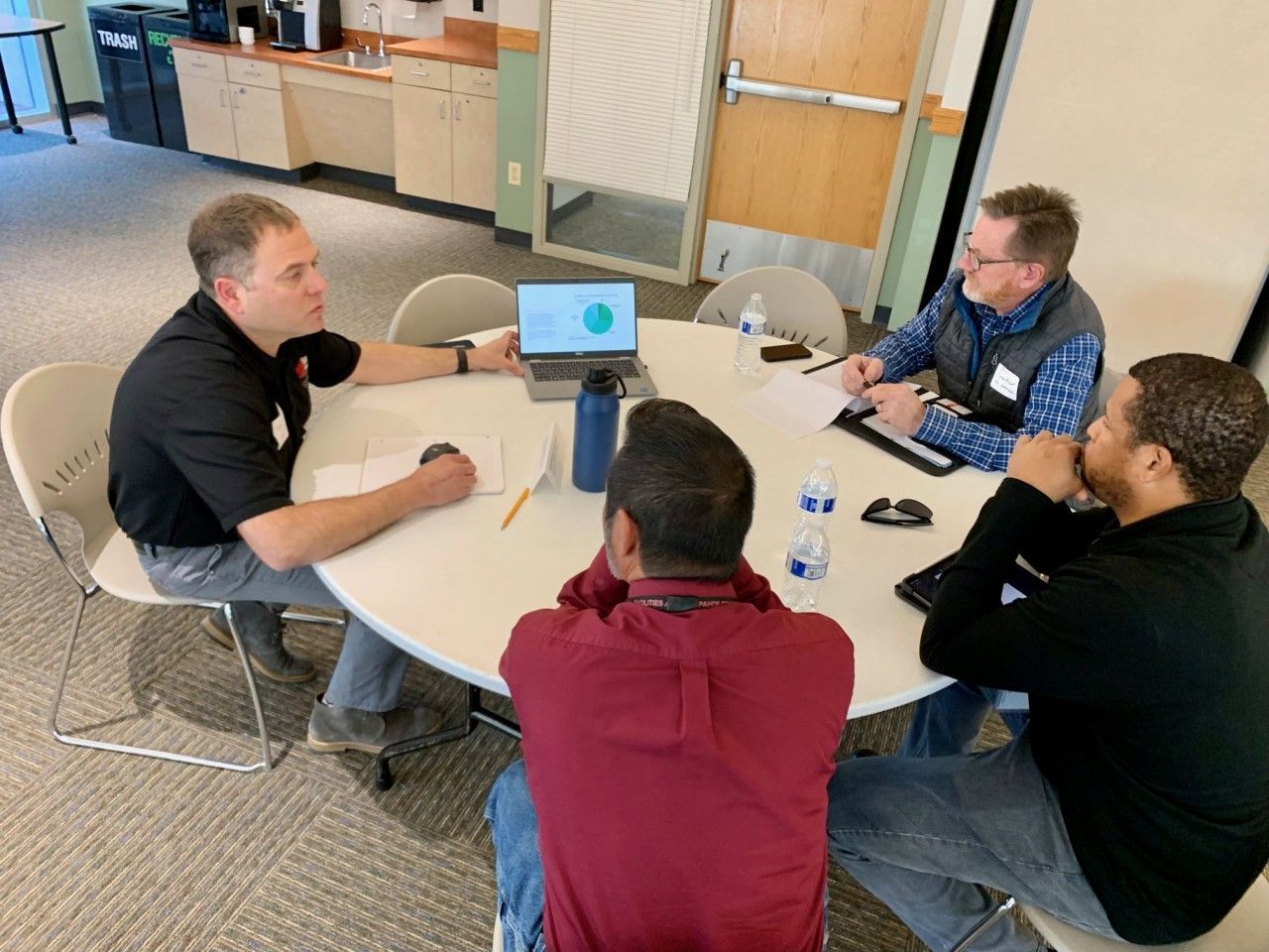 Four people seated around a white table, looking at a laptop and documents, discussing.