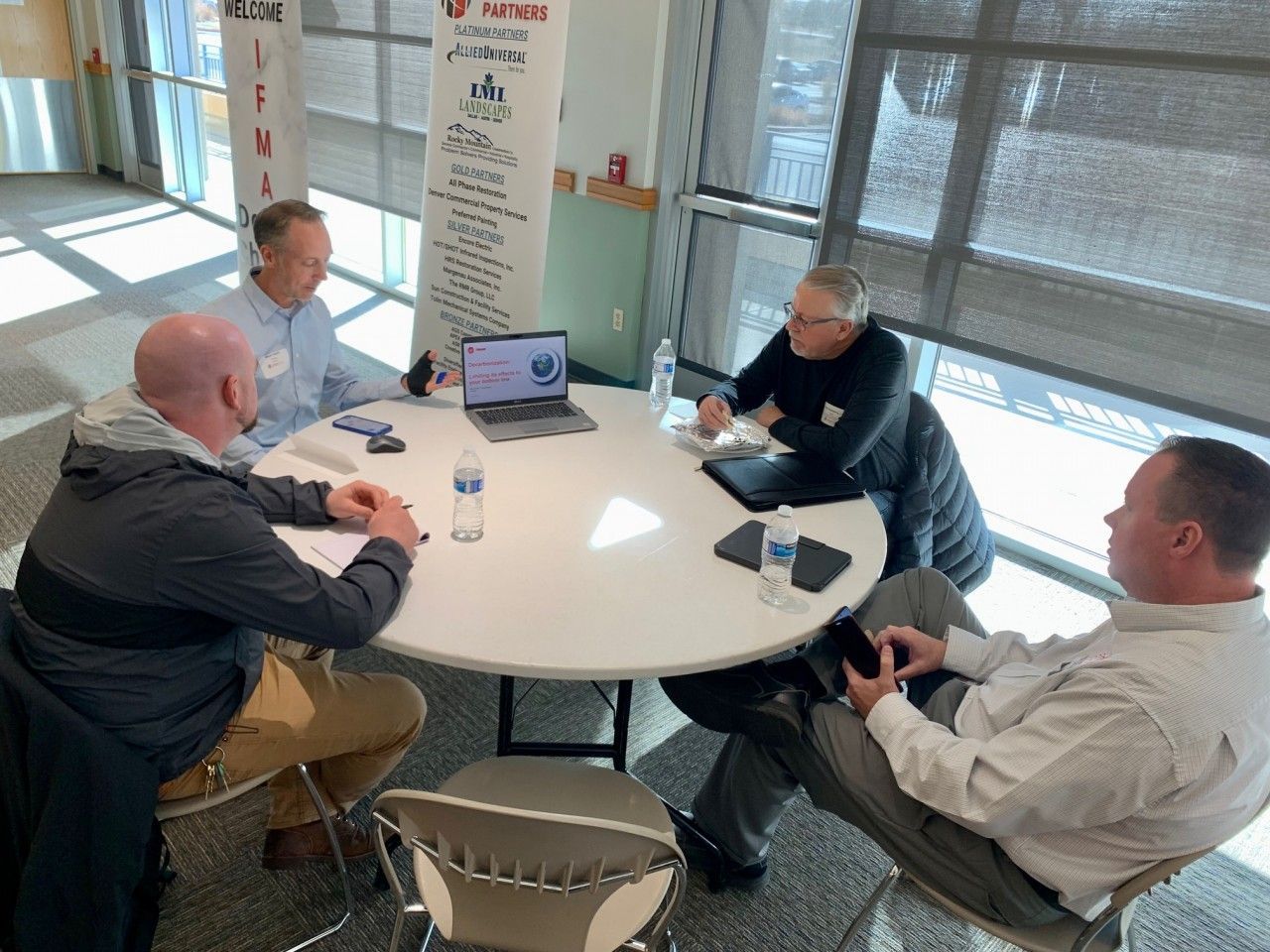 Four people seated around a round table, discussing; a laptop sits on the table; sunny interior.