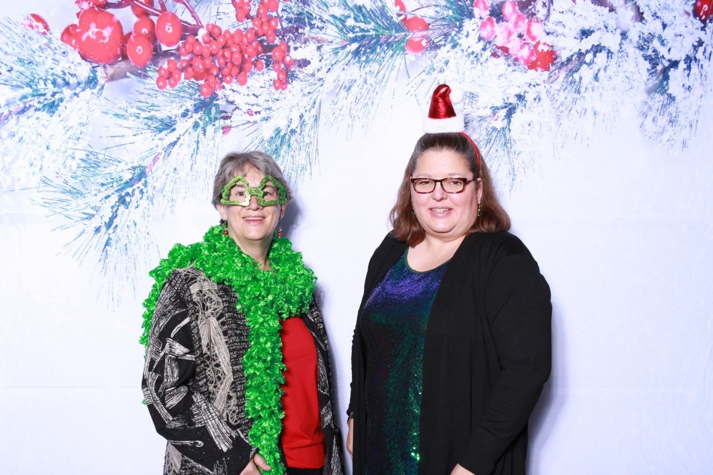 Two people pose in front of a festive backdrop. One wears green boa and mask; the other, glasses and a mini Santa hat.
