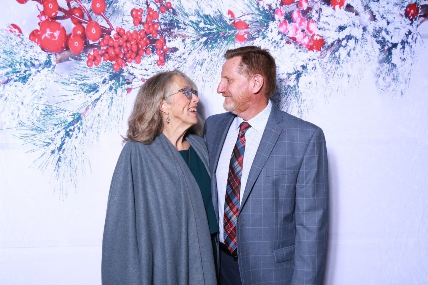 Couple smiling at each other, posing in front of a holiday-themed backdrop of berries and snowy branches.