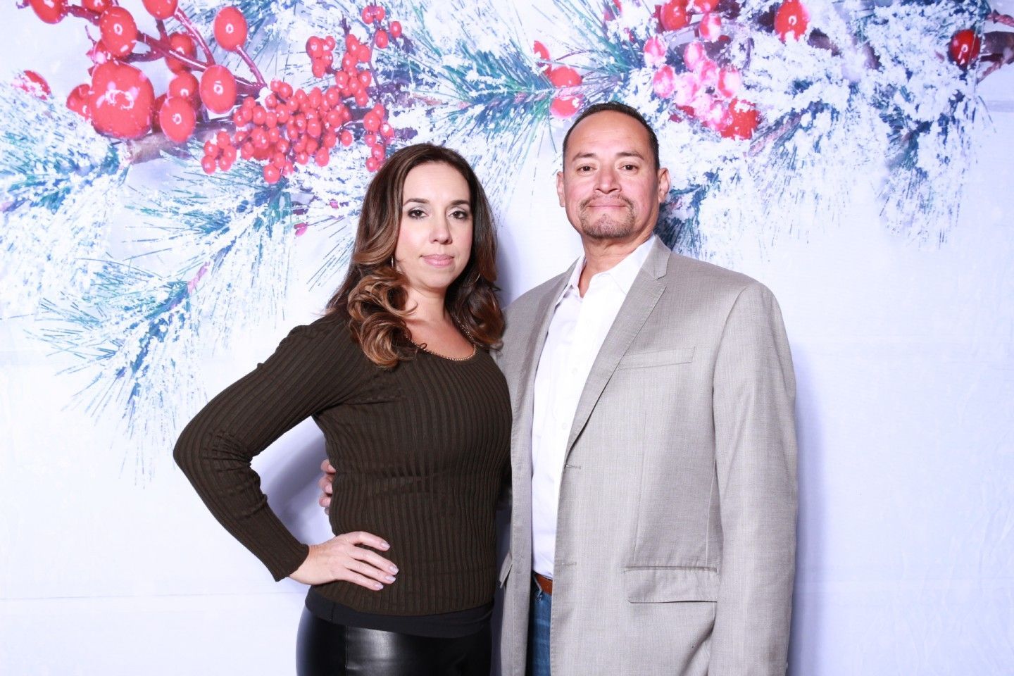 Couple posing in front of a holiday-themed backdrop with berries and snowy branches. The woman has her hand on her hip.