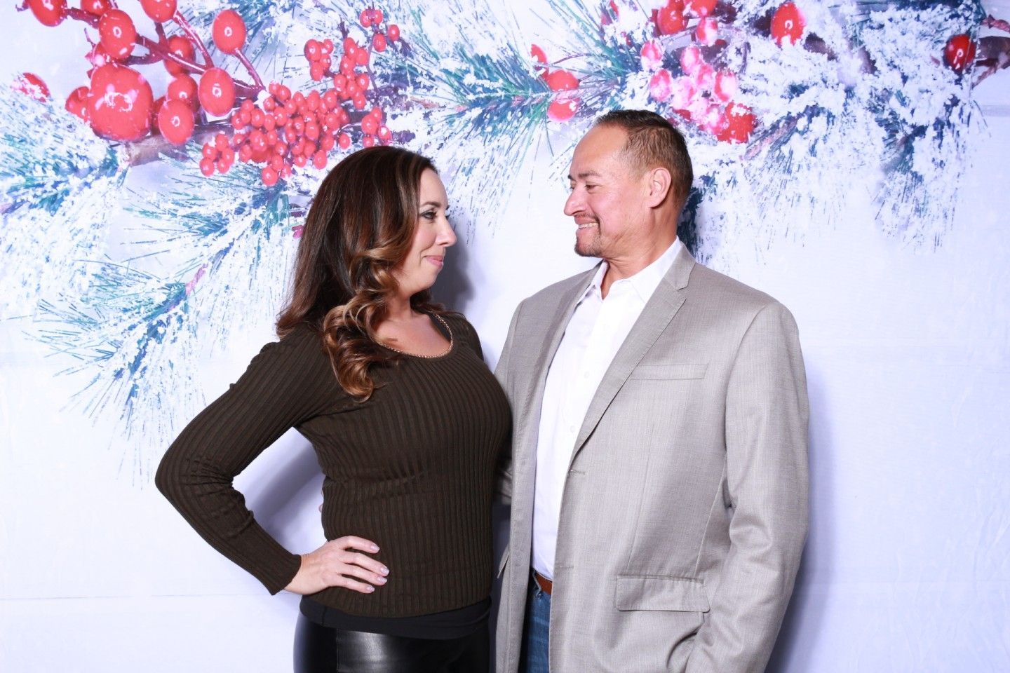 Couple smiling, posing in front of a holiday-themed backdrop of berries and snowy branches.