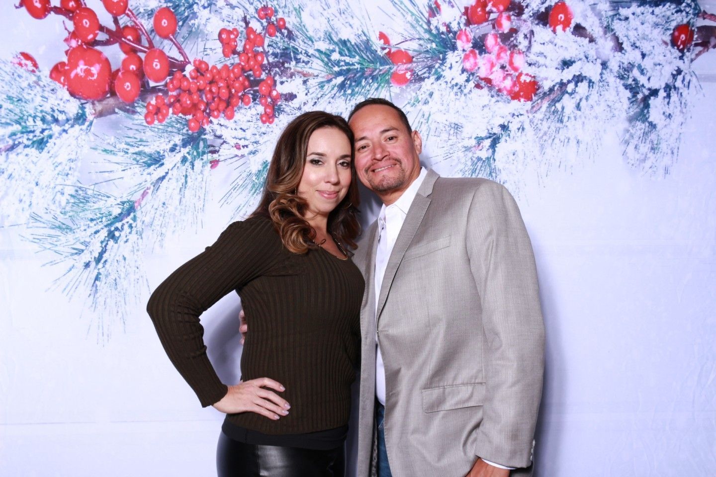 Couple posing in front of holiday backdrop with red berries and snowy branches. Woman in brown sweater, man in blazer.
