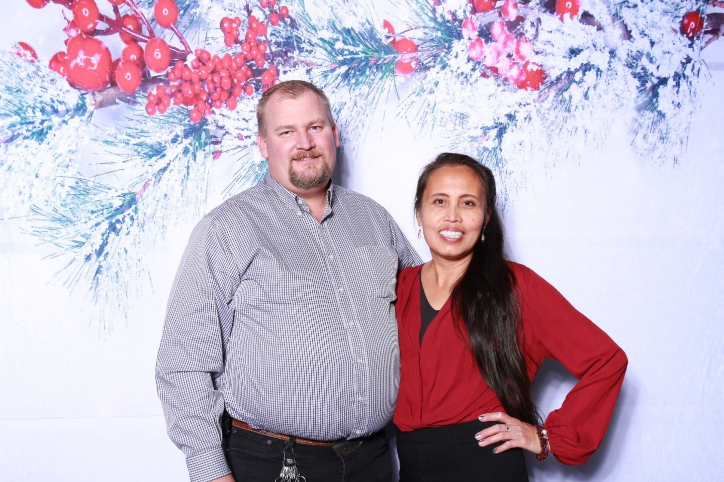 Man and woman posing in front of a snowy, red berry background. They are smiling.