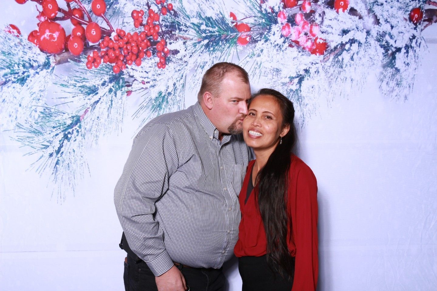 Man kissing woman's cheek; couple posing in front of festive backdrop with red berries and snow-covered branches.