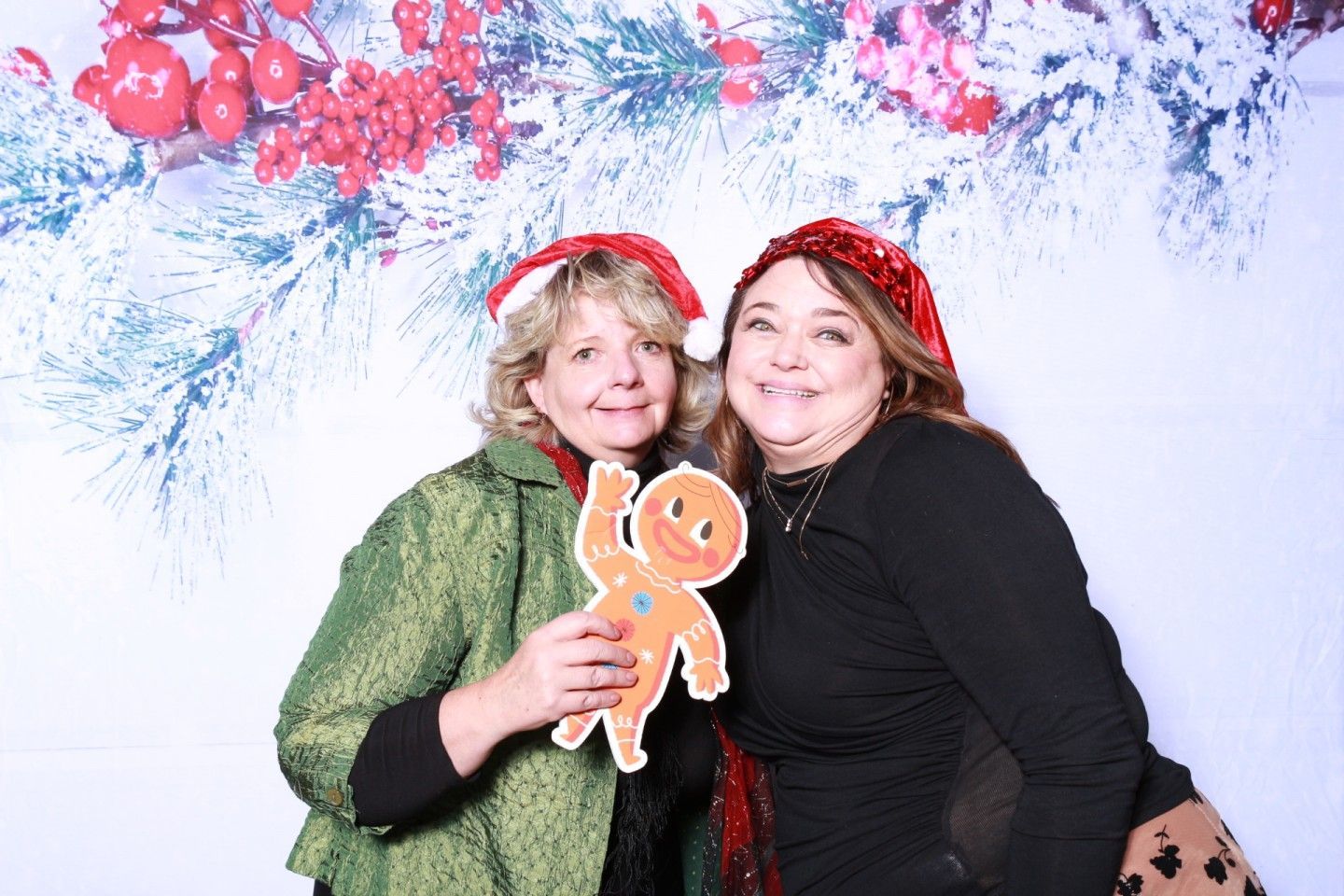 Two women pose with a gingerbread cutout against a holiday backdrop; one woman is holding the cutout.