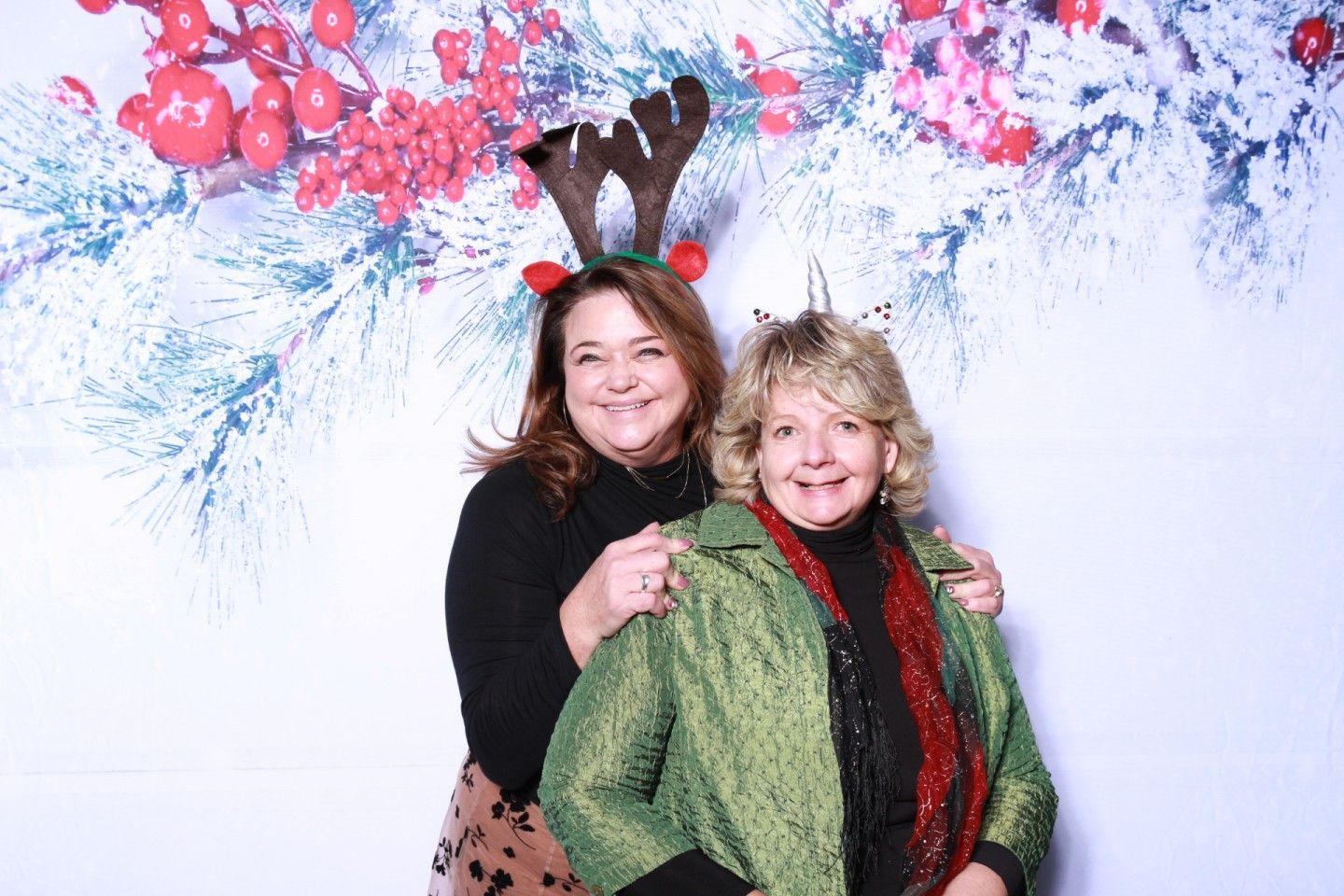 Two women pose for a photo in front of a holiday backdrop. One wears reindeer antlers.