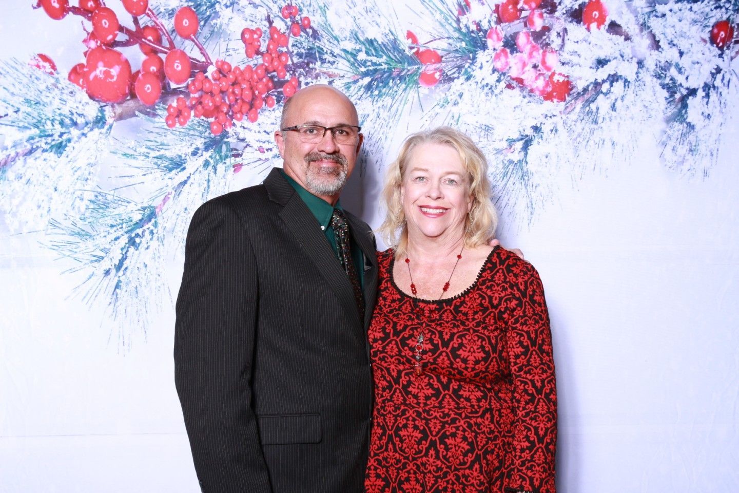 Couple poses in front of a holiday backdrop with red berries and snow-covered branches.