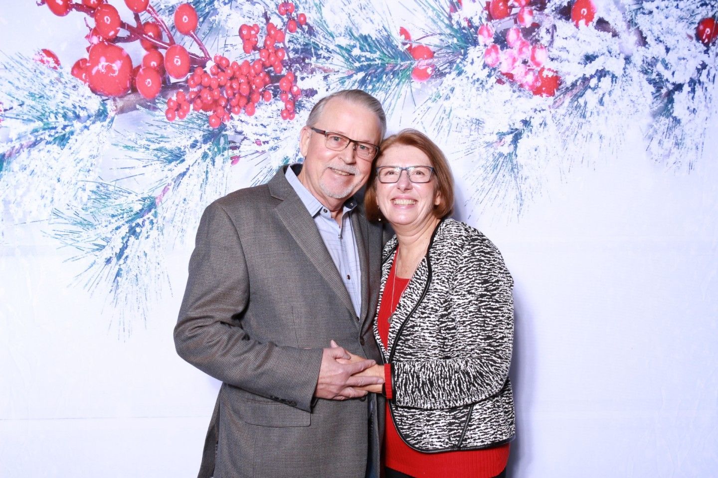 Couple posing, smiling, holding hands, in front of a holiday-themed backdrop with red berries and snow.