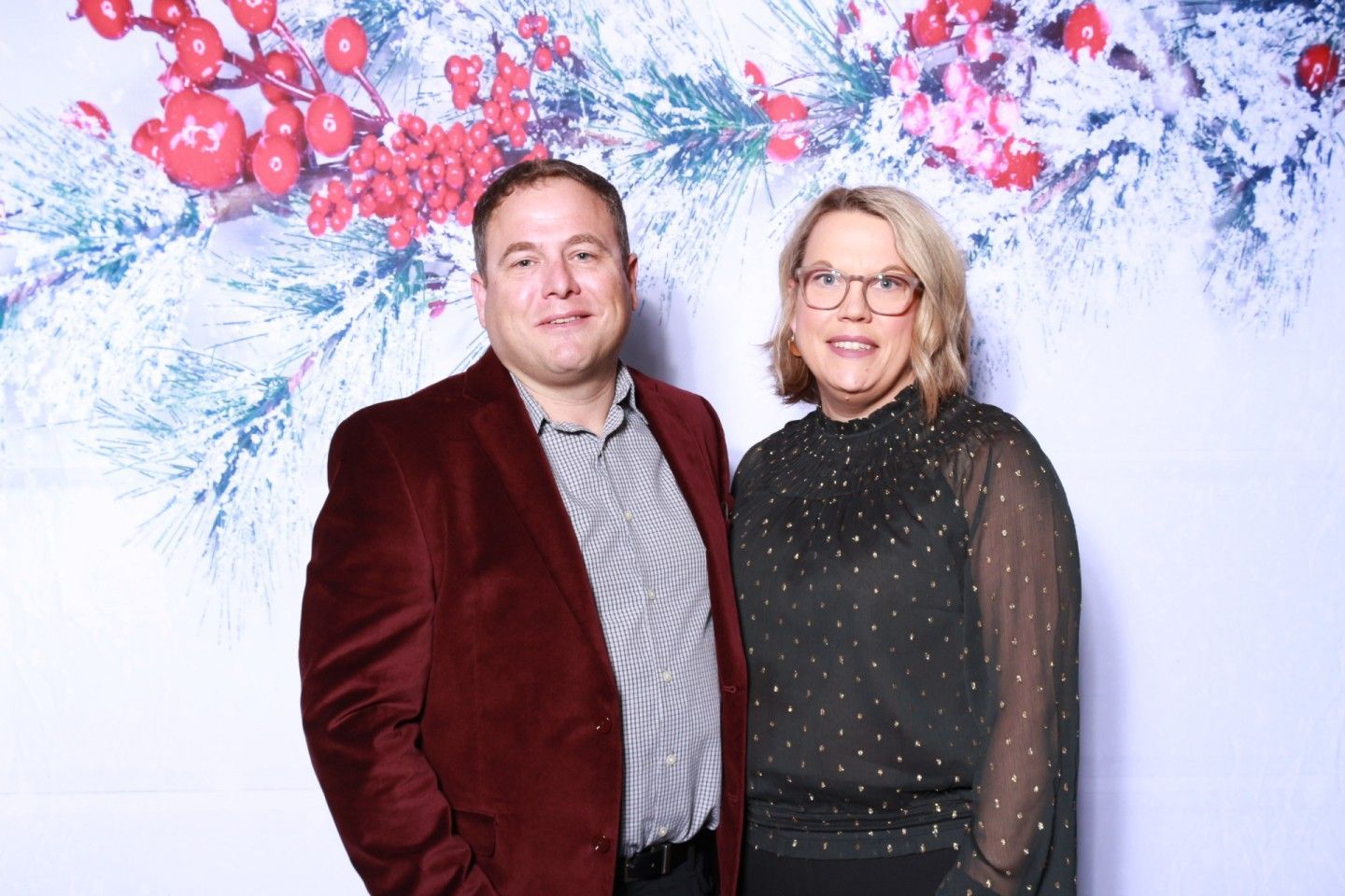 Man in red velvet jacket and woman in black top pose in front of a holiday backdrop.