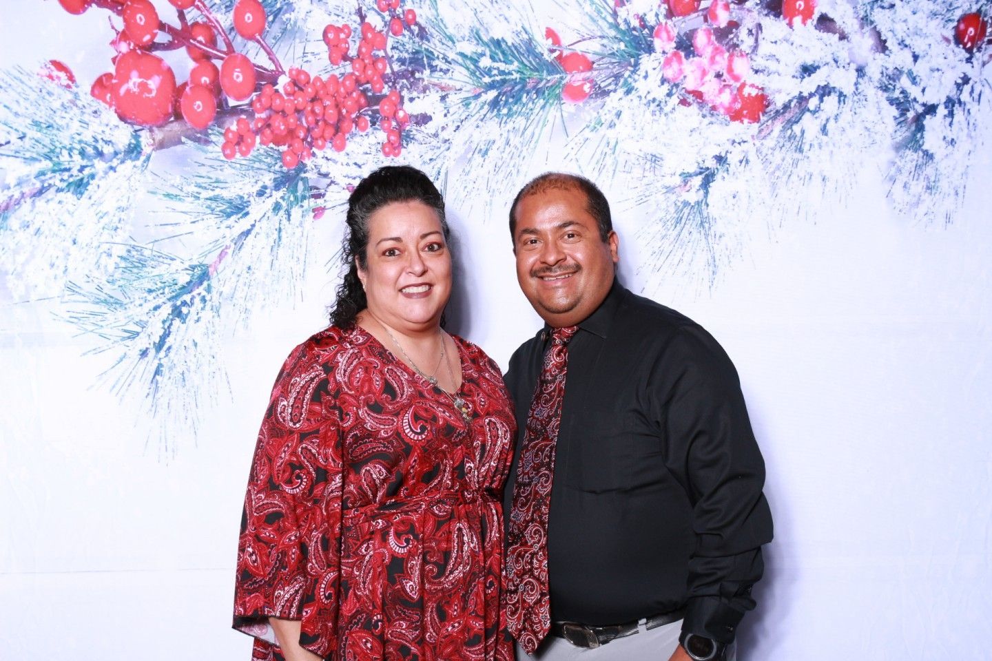 Couple posing in front of a holiday backdrop. Woman wears a red dress, man in black shirt and tie.