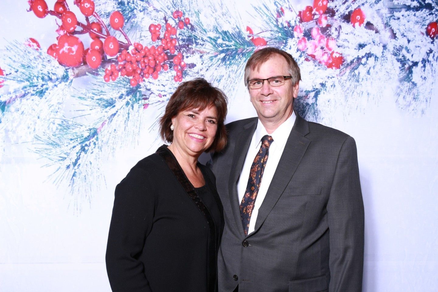 Man and woman posing, smiling, against a winter-themed backdrop with red berries and greenery.