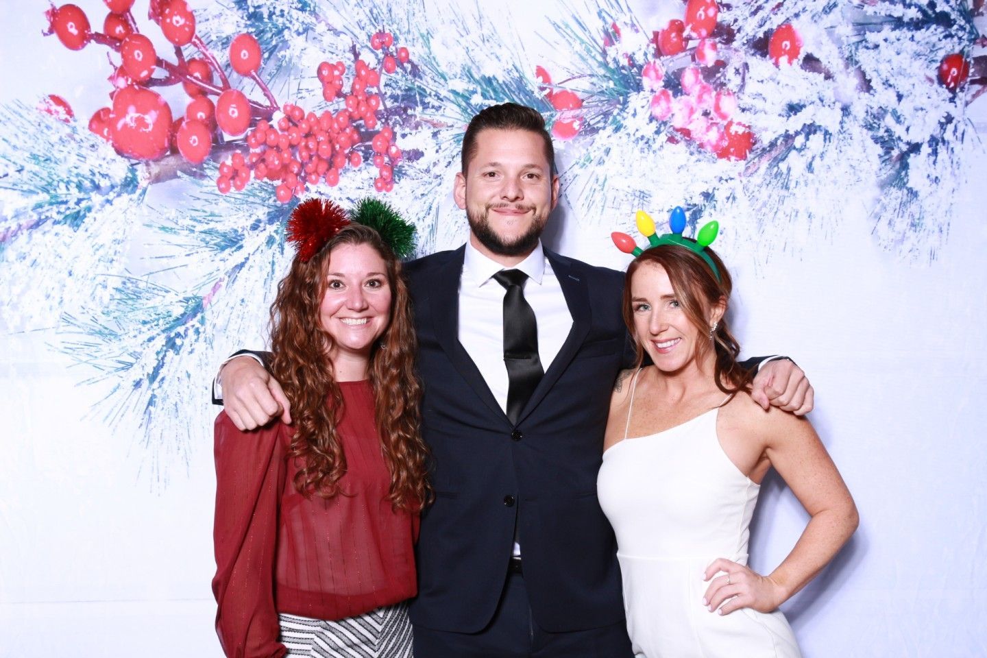Three people posing for photo: man in suit, two women with holiday headbands, winter backdrop.