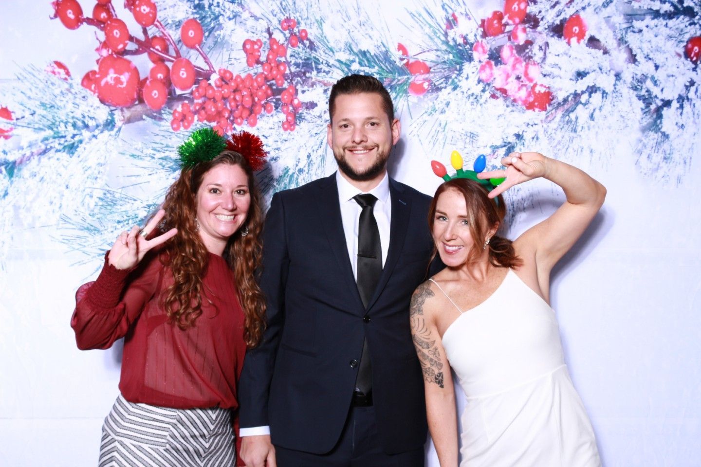 Three people pose in front of a holiday backdrop. A man in a suit stands between two women wearing festive headbands.