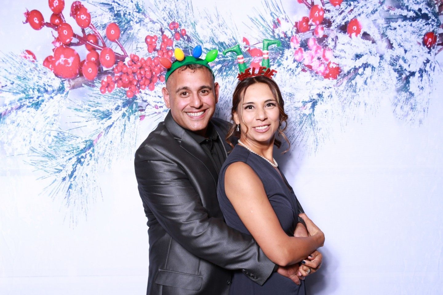 Couple smiling, hugging in front of winter berry backdrop, wearing festive headbands.