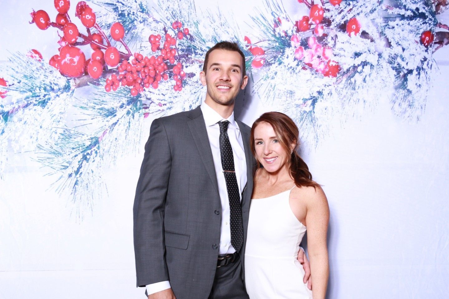 Couple smiling in front of a white backdrop with red berries and snowy branches. Man in suit, woman in white dress.