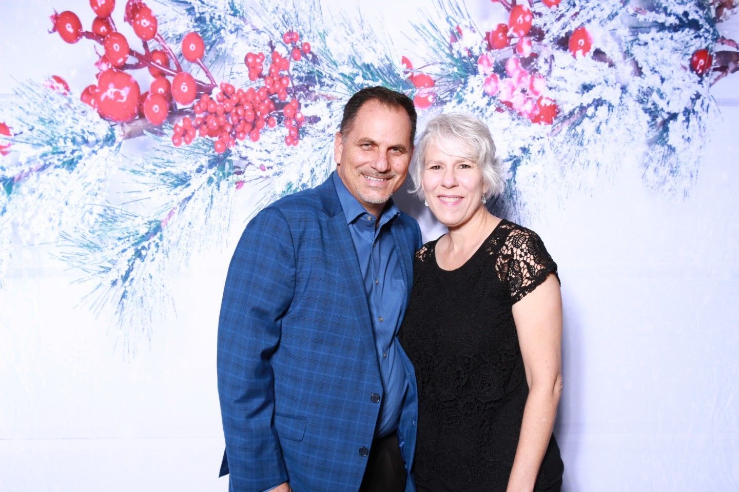 Man and woman smiling, posing in front of festive winter backdrop. Man in blue jacket, woman in black dress.