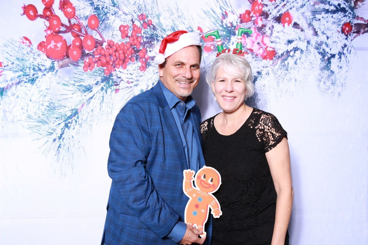 Couple poses in front of a holiday backdrop. Man holds a gingerbread cutout, both smiling, festive hats.