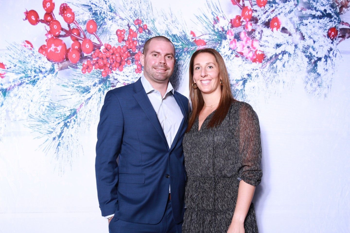 A man and woman pose in front of a holiday-themed backdrop. They smile at the camera.