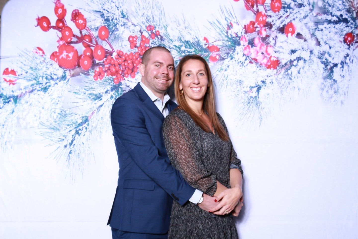 Couple poses in front of a holiday backdrop with berry and snow design. The man hugs the woman.