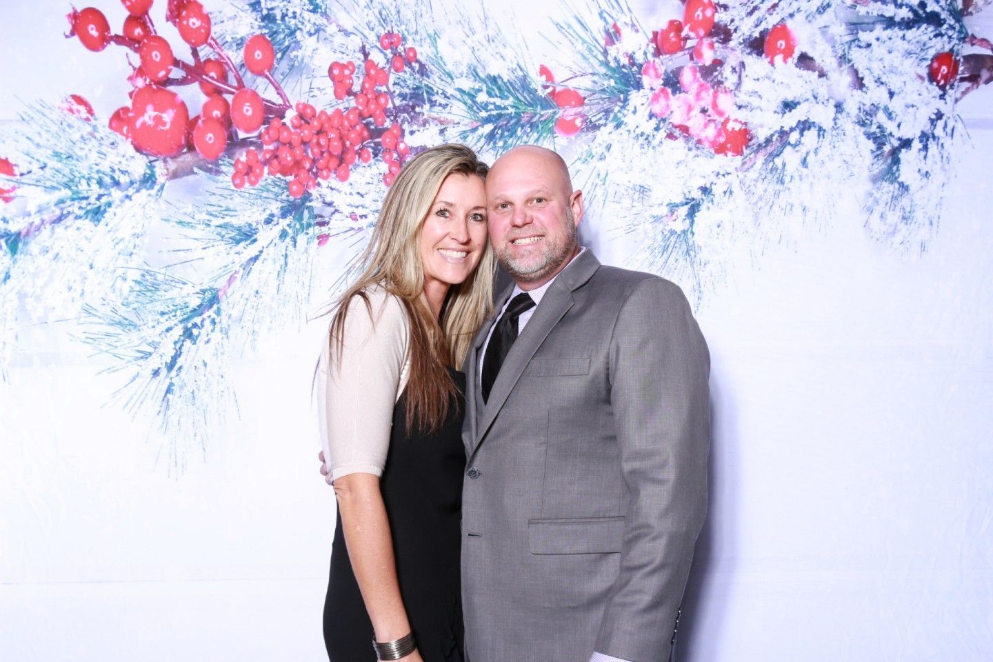 Couple smiling, posing in front of a holiday backdrop of snow-covered branches and red berries.