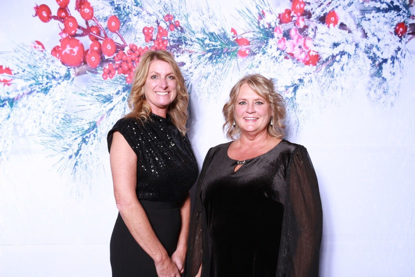 Two women pose in front of a holiday-themed backdrop. One wears a black sequin top, the other a black dress.
