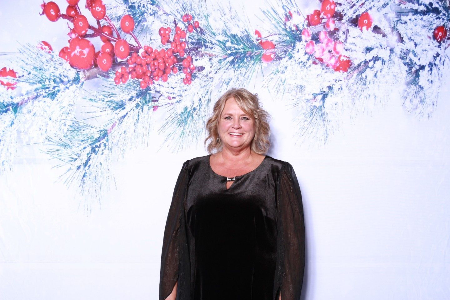 Woman in a black dress smiling in front of a white backdrop with winter greenery and red berries.