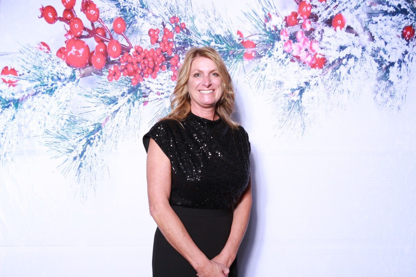 Woman in black dress smiling, posing in front of white backdrop with red berries and pine boughs.